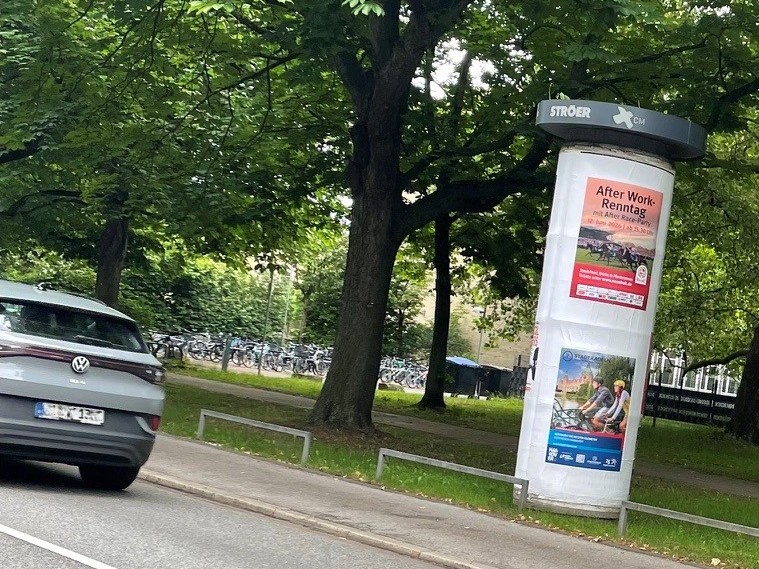 Eine Litfasssäule steht am Straßenrand und ist mit einem A0-Affichenplakat in der oberen Säulenhälfte plakatiert.